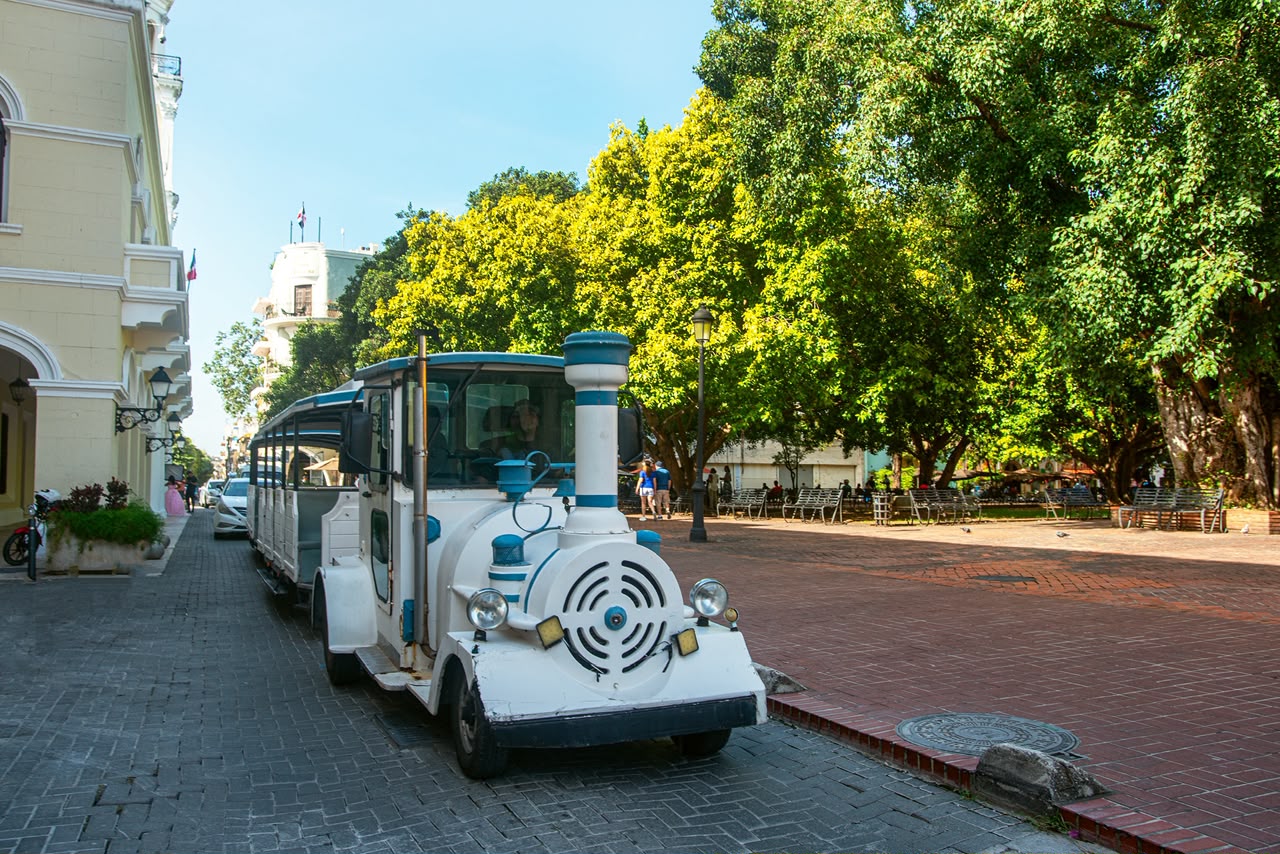 Tren turístico junto a un parque con grandes árboles en la Zona Colonial