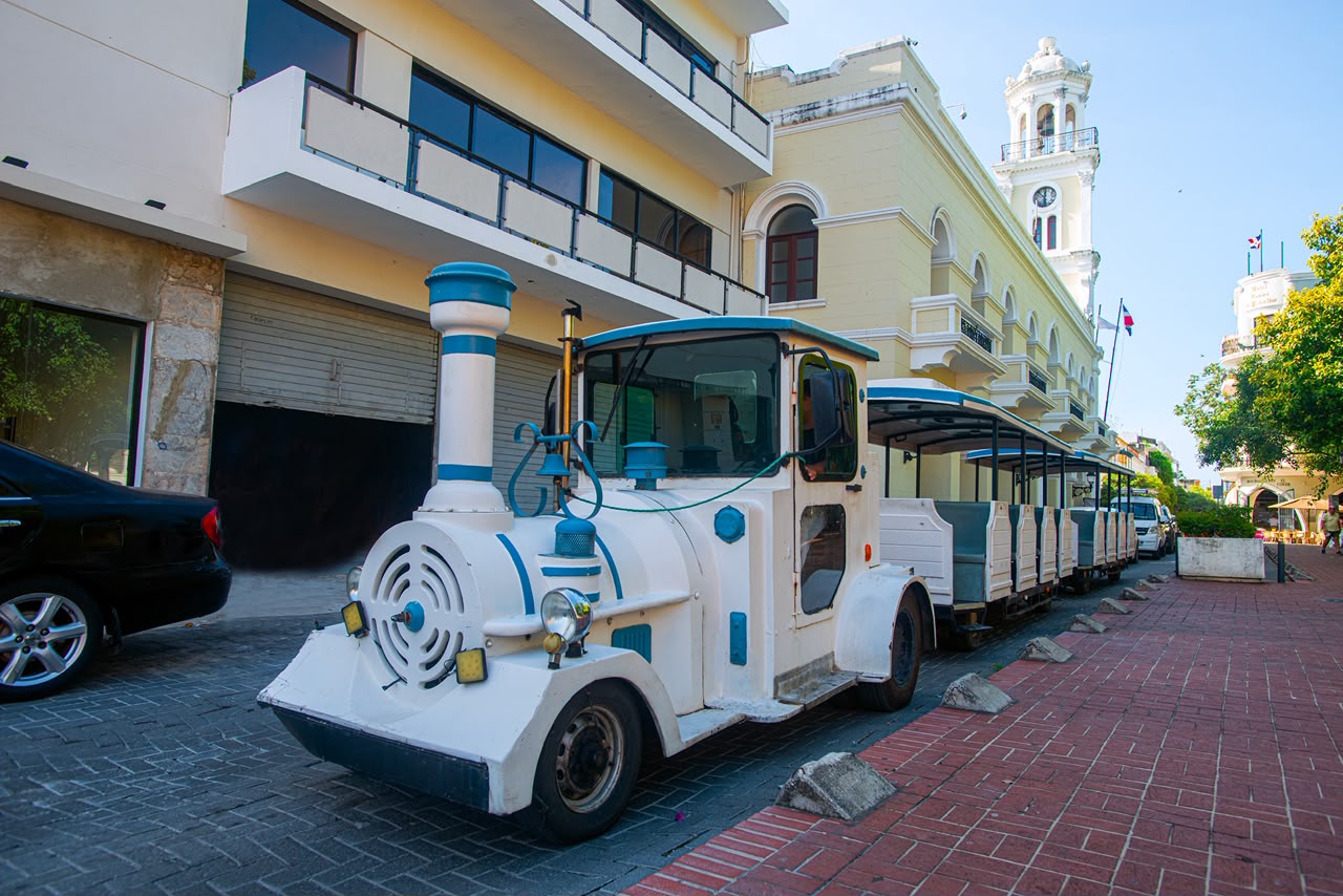 Tren turístico junto a un edificio moderno con torre colonial de fondo